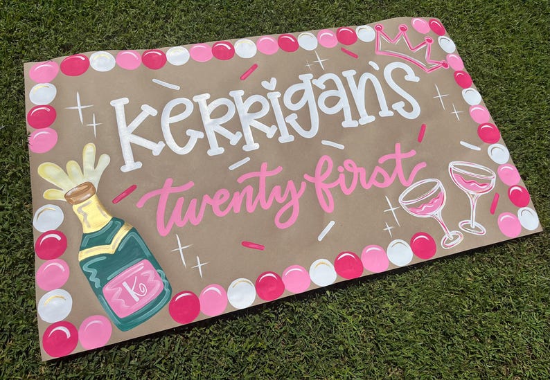 May include: A hand-painted sign on brown paper, celebrating a twenty-first birthday. The sign features the name "Kerrigans" in white, and "twenty first" in pink. It includes a champagne bottle, two wine glasses, and a crown.