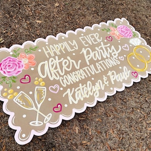 May include: A decorative sign with a scalloped edge, featuring the words "Happily Ever After Party Congratulations Katelyn & Paul" in white script. The sign includes floral and champagne glass illustrations, along with hearts and wedding rings.
