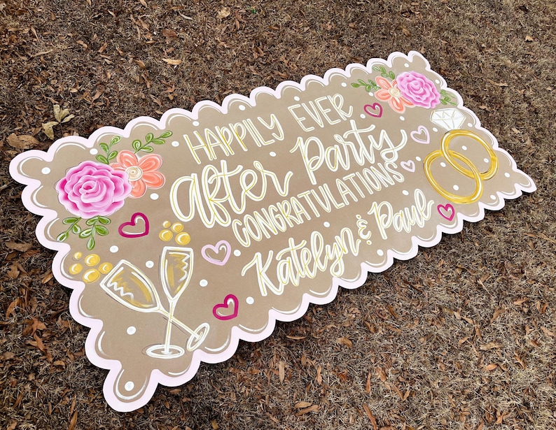 May include: A decorative sign with a scalloped edge, featuring the words "Happily Ever After Party Congratulations Katelyn & Paul" in white script. The sign includes floral and champagne glass illustrations, along with hearts and wedding rings.