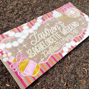 May include: A hand-painted sign on brown paper with the text "Lauren's BACHELORETTE WEEKEND Dallas TX May 7-10". The sign features illustrations of a diamond ring, champagne bottle, and pearl necklace, with pink and orange accents.
