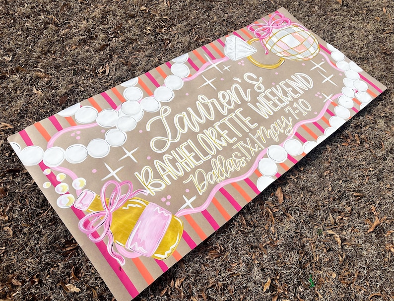 May include: A hand-painted sign on brown paper with the text "Lauren's BACHELORETTE WEEKEND Dallas TX May 7-10". The sign features illustrations of a diamond ring, champagne bottle, and pearl necklace, with pink and orange accents.