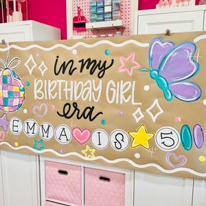 May include: A brown banner with the text "in my BIRTHDAY GIRL era EMMA IS 5" and colorful illustrations, including a disco ball, butterfly, stars, and hearts. The banner is decorated with a white border.