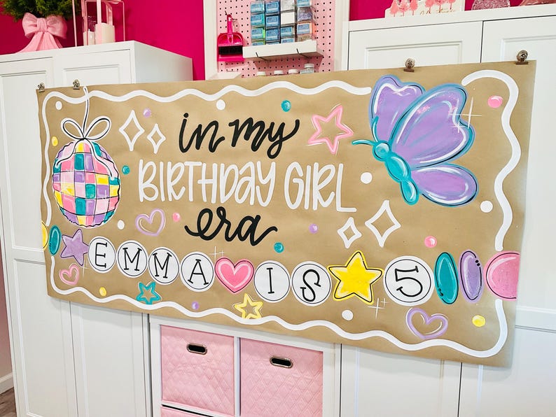 May include: A brown banner with the text "in my BIRTHDAY GIRL era EMMA IS 5" and colorful illustrations, including a disco ball, butterfly, stars, and hearts. The banner is decorated with a white border.