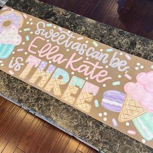 May include: A hand-painted banner with the text "Sweet as can be Ella Kate is THREE." The banner features illustrations of donuts, cupcakes, ice cream cones, and macarons in pastel colors. Sprinkles and dots are scattered around the text and illustrations.