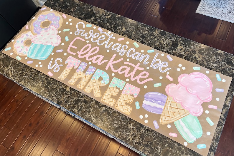 May include: A hand-painted banner with the text "Sweet as can be Ella Kate is THREE." The banner features illustrations of donuts, cupcakes, ice cream cones, and macarons in pastel colors. Sprinkles and dots are scattered around the text and illustrations.