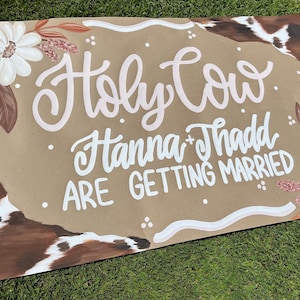 May include: A rustic wedding sign with the phrase "Holy Cow Hanna + Thadd ARE GETTING MARRIED" in white script. The sign features floral and cowhide accents, with white daisies and brown leaves. The background is a tan color.