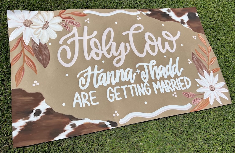May include: A rustic wedding sign with the phrase "Holy Cow Hanna + Thadd ARE GETTING MARRIED" in white script. The sign features floral and cowhide accents, with white daisies and brown leaves. The background is a tan color.