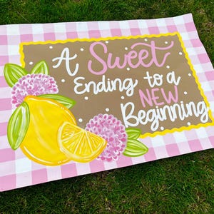 May include: A decorative sign with a pink and white gingham pattern border. The sign features a brown center with the text "A Sweet Ending to a NEW Beginning" in white and pink script. A yellow lemon and pink flowers are painted on the sign.
