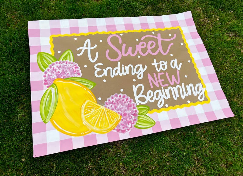 May include: A decorative sign with a pink and white gingham pattern border. The sign features a brown center with the text "A Sweet Ending to a NEW Beginning" in white and pink script. A yellow lemon and pink flowers are painted on the sign.