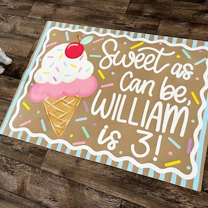 May include: A rectangular sign with a brown background features a large ice cream cone illustration. The sign reads "Sweet as Can be, WILLIAM is 3!" in white script. The ice cream has pink, white, and red accents.