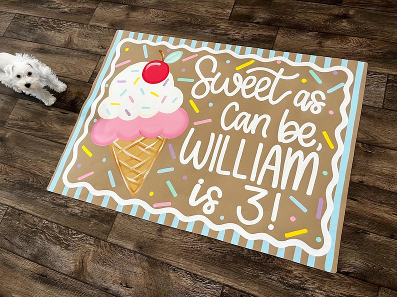 May include: A rectangular sign with a brown background features a large ice cream cone illustration. The sign reads "Sweet as Can be, WILLIAM is 3!" in white script. The ice cream has pink, white, and red accents.