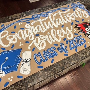 May include: A graduation sign with the words "Congratulations Briley Class of 2025" in white and blue. The sign includes a graduation cap, diploma, and a "SEMO Bound" logo. The background is tan.