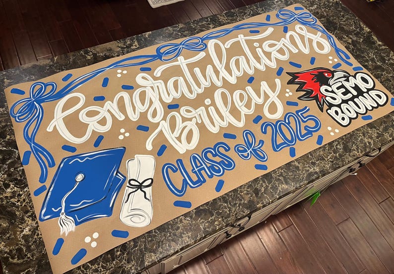May include: A graduation sign with the words "Congratulations Briley Class of 2025" in white and blue. The sign includes a graduation cap, diploma, and a "SEMO Bound" logo. The background is tan.