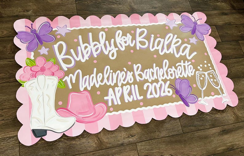 May include: A decorative sign with a scalloped pink border and a brown center. The sign features the text "Bubbly for Bialka Madeline's Bachelorette APRIL 2026" in white script, along with illustrations of a cowboy boot, a cowboy hat, butterflies, and champagne glasses.