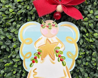 May include: A wooden angel ornament with a red bow and bead accents. The angel has a white dress, gold star, and blue and gold wings. The ornament is decorated with holly and berries, perfect for Christmas.