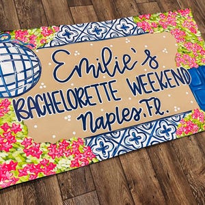 May include: A decorative mat with a disco ball and floral design. The mat features the text "Emilie's BACHELORETTE WEEKEND Naples, FL." in blue script on a tan background, surrounded by pink flowers and blue accents.
