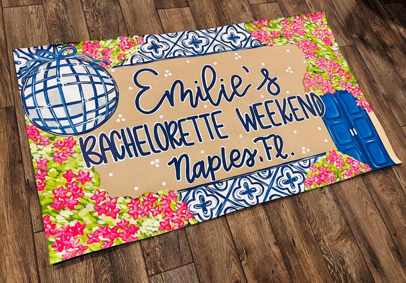 May include: A decorative mat with a disco ball and floral design. The mat features the text "Emilie's BACHELORETTE WEEKEND Naples, FL." in blue script on a tan background, surrounded by pink flowers and blue accents.