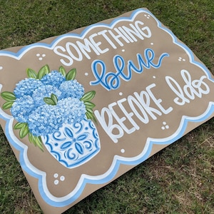May include: A brown sign with the text "SOMETHING blue BEFORE I do" in white and blue lettering. A blue and white vase with blue flowers is on the left side. The sign has a scalloped edge with a blue border.