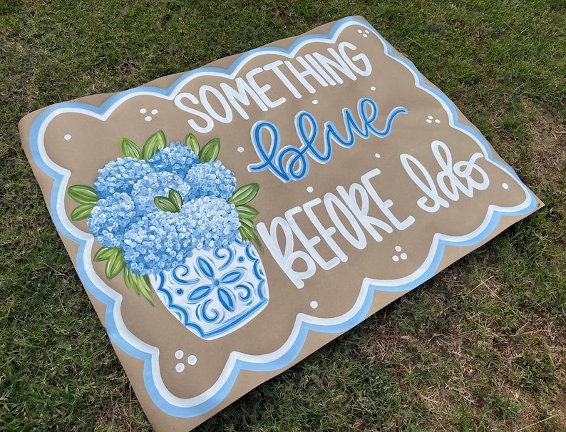 May include: A brown sign with the text "SOMETHING blue BEFORE I do" in white and blue lettering. A blue and white vase with blue flowers is on the left side. The sign has a scalloped edge with a blue border.