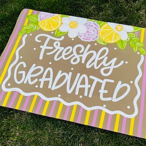 May include: A rectangular sign with the words "Freshly GRADUATED" in white script. The sign has a brown background with a white scalloped border. It is decorated with yellow lemons, white flowers, and pink and yellow stripes.