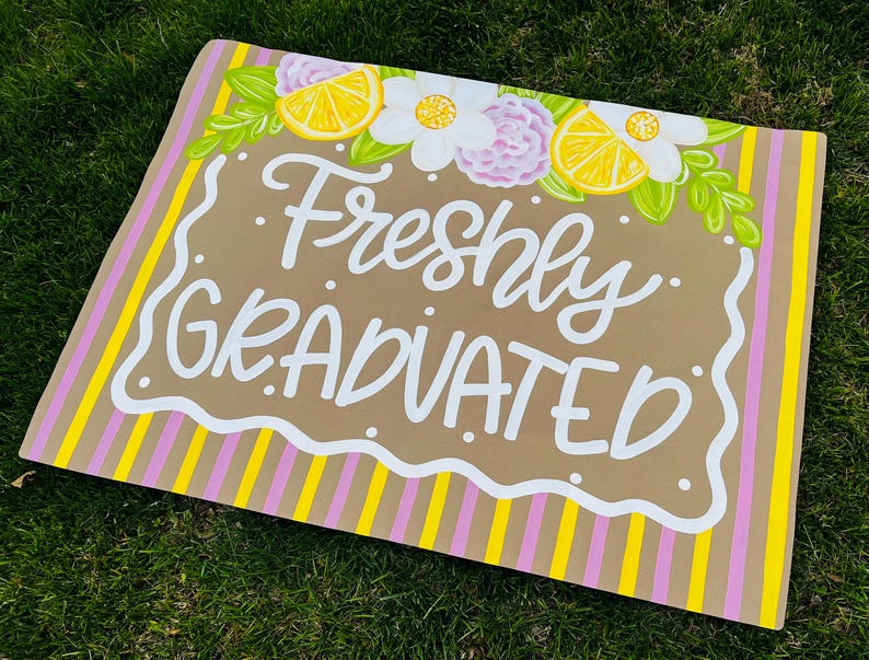 May include: A rectangular sign with the words "Freshly GRADUATED" in white script. The sign has a brown background with a white scalloped border. It is decorated with yellow lemons, white flowers, and pink and yellow stripes.