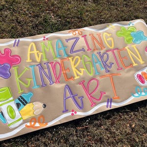 May include: A brown paper banner with the words "AMAZING KINDERGARTEN ART" in colorful bubble letters. The banner features paint splatters, a paintbrush, crayons, and a pencil.
