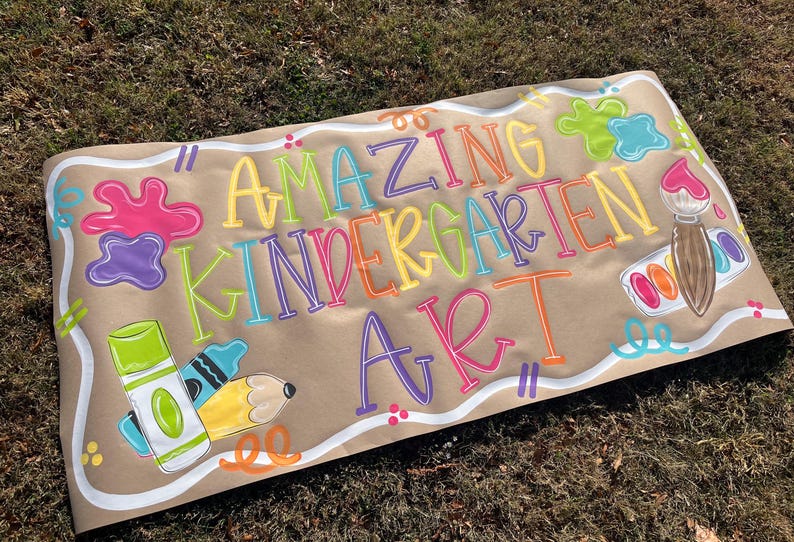 May include: A brown paper banner with the words "AMAZING KINDERGARTEN ART" in colorful bubble letters. The banner features paint splatters, a paintbrush, crayons, and a pencil.