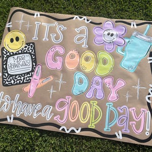 May include: A hand-painted sign on brown paper with the words "It's a Good Day to have a Good Day!" in colorful bubble letters. Includes a smiley face, flower, and drink illustration.