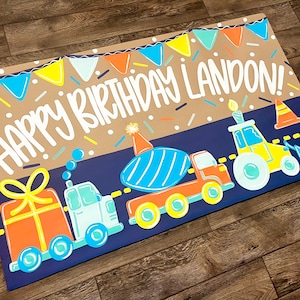 May include: A colorful birthday-themed illustration featuring the text "HAPPY BIRTHDAY LANDON!" above a row of cartoon vehicles. The design includes a gift, a truck, a cement mixer, and a tractor, all in bright colors, with confetti and bunting.