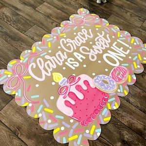 May include: A decorative sign with a scalloped edge, featuring the text "Clara Grace is a Sweet ONE!" in pink. The sign includes a pink cake with a candle, donuts, and colorful sprinkles. The background is a light brown color.