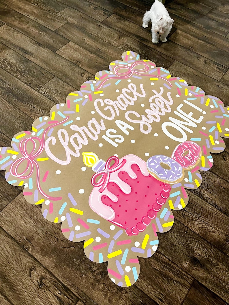 May include: A decorative sign with a scalloped edge, featuring the text "Clara Grace is a Sweet ONE!" in pink. The sign includes a pink cake with a candle, donuts, and colorful sprinkles. The background is a light brown color.