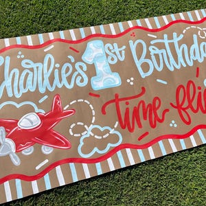 May include: A brown banner with the text "Charlie's 1st Birthday Time flies" in blue and red lettering. A red airplane graphic is on the left. The banner has a red and white striped border.