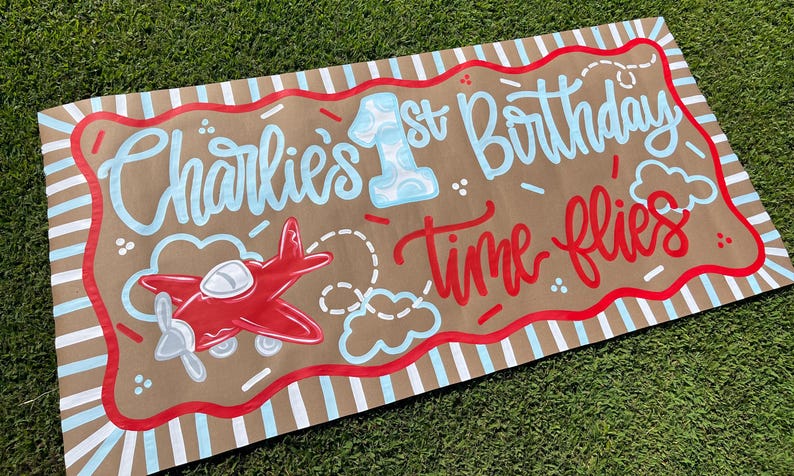 May include: A brown banner with the text "Charlie's 1st Birthday Time flies" in blue and red lettering. A red airplane graphic is on the left. The banner has a red and white striped border.