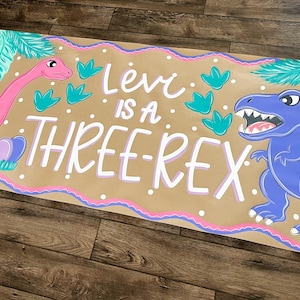 May include: A hand-painted banner with a brown background, featuring a pink dinosaur, a blue dinosaur, and the text "Levi is a THREE-REX." The banner is decorated with teal leaves and a pink and purple border.