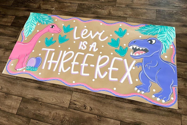 May include: A hand-painted banner with a brown background, featuring a pink dinosaur, a blue dinosaur, and the text "Levi is a THREE-REX." The banner is decorated with teal leaves and a pink and purple border.