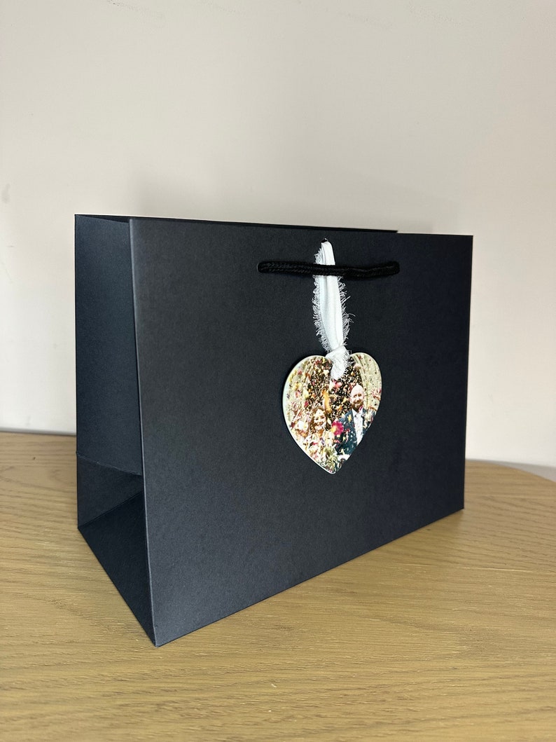 Peut inclure: Un sac cadeau noir avec une &eacute;tiquette photo en forme de c&oelig;ur attach&eacute;e. L'&eacute;tiquette photo pr&eacute;sente un groupe de personnes dans un cadre de mariage.