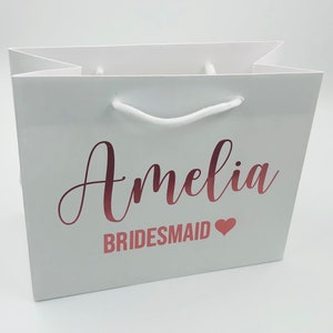 May include: White gift bag with a rose gold foil design. The bag says "Amelia Bridesmaid" with a heart.