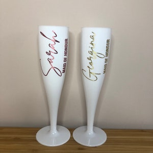 May include: Two white plastic champagne flutes with gold and pink lettering. The left flute says "Sarah Maid of Honour" and the right flute says "Georgina Maid of Honour".