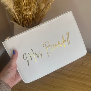 May include: White clutch bag with a gold zipper and the text "Mrs. Beardall" written in gold script.