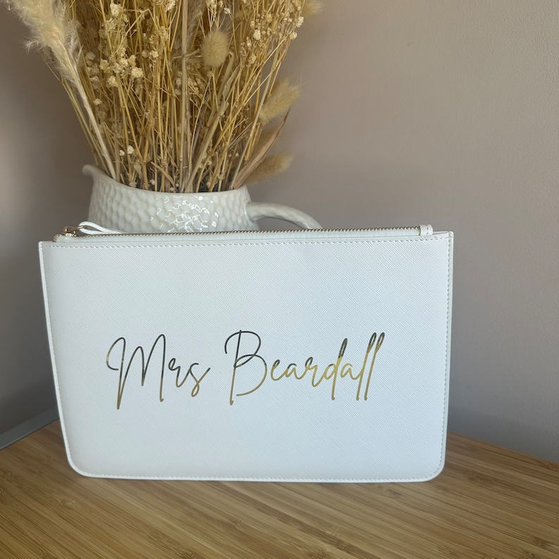 May include: White zippered clutch bag with a gold inscription that reads "Mrs. Beardall".