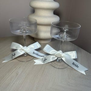 May include: Two clear glass champagne flutes with white satin ribbons tied around the stems. The ribbons are labelled "Bride", "Maid of Honour", and "Bridesmaid".