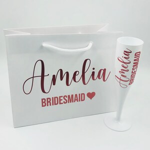 May include: White gift bag with a white rope handle. The bag has the text "Amelia Bridesmaid" in rose gold lettering with a small pink heart. A white plastic champagne flute with the text "Amelia Bridesmaid" in rose gold lettering is next to the bag.