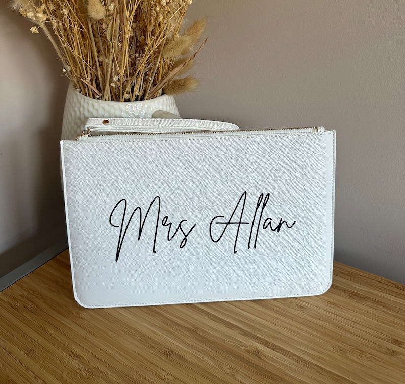 May include: White clutch bag with a zip closure. The bag has the text "Mrs Allan" printed in black cursive script.