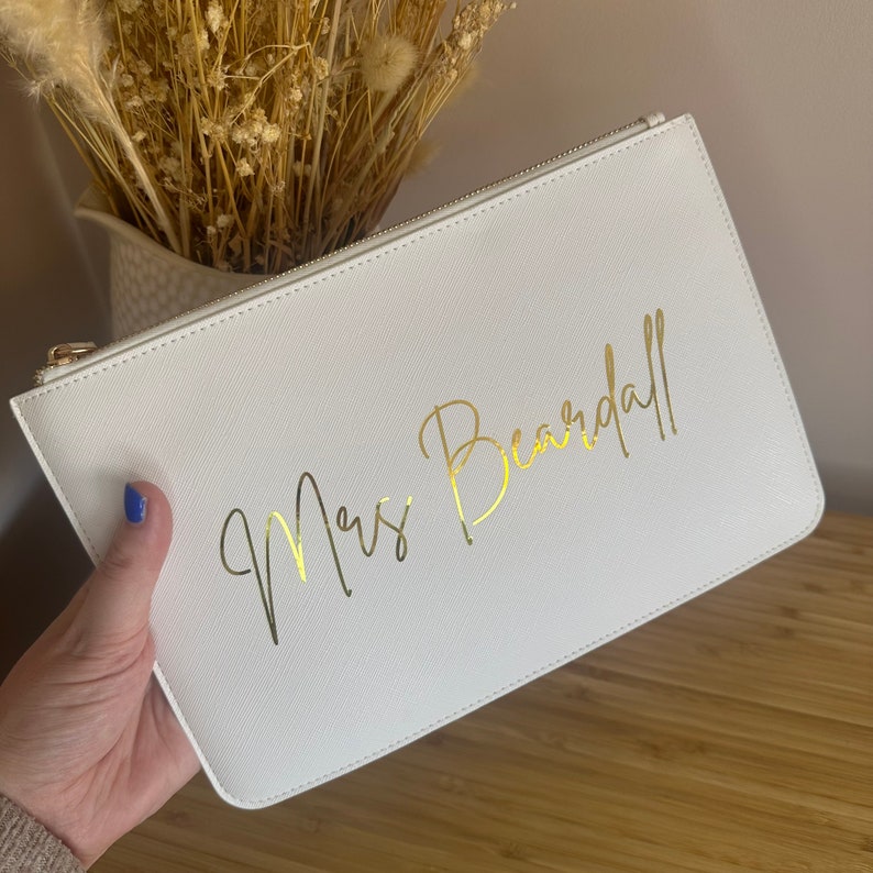 May include: White clutch bag with a gold zipper and the text "Mrs. Beardall" written in gold script.