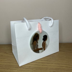 Peut inclure: Un sac cadeau blanc avec une photo ronde de deux personnes en tenue de mariage attach&eacute;e au sac avec un ruban rose.