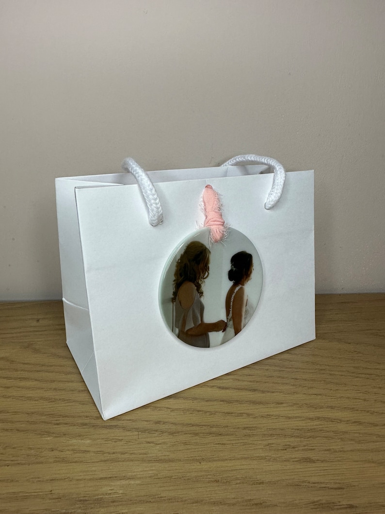 Peut inclure: Un sac cadeau blanc avec une photo ronde de deux personnes en tenue de mariage attach&eacute;e au sac avec un ruban rose.