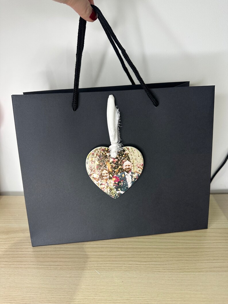 Peut inclure: Un sac cadeau noir avec une anse en cordon blanc. Un ornement en forme de c&oelig;ur avec une photo d'un groupe de mariage est attach&eacute; &agrave; l'anse.