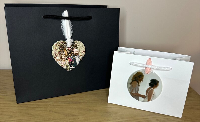 Peut inclure: Deux sacs cadeaux blancs avec des poign&eacute;es en corde noire et blanche. Un sac a un ornement photo en forme de c&oelig;ur avec une photo d'un groupe de personnes. L'autre sac a un ornement photo rond avec une photo de deux personnes.