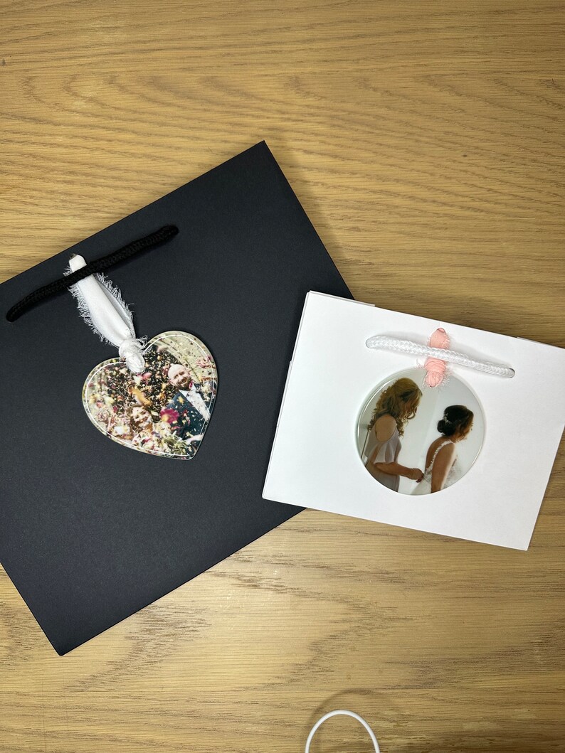 Peut inclure: Deux sacs cadeaux blancs avec des poign&eacute;es en ruban noir et rose. Un sac a une photo en forme de c&oelig;ur d'un couple devant un fond de confettis. L'autre sac a une photo ronde de deux personnes en robe blanche et en robe blanche.