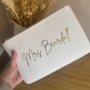 May include: White clutch bag with a gold zipper and the text "Mrs. Beardall" written in gold script.